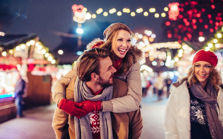 Young couple having fun outdoors at winter fair. Wearing warm clothes, hats and scarfs. Visiting christmas market in Vienna, Austria. Everything is decorated with festive string lights. Evening or night with beautiful lights lightning the scenes.