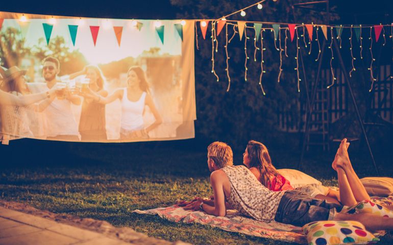 Young couple having movie night party. Laying down on blanket in front of movie improvised screen. Backyard decorated with festive string lights. Night time.