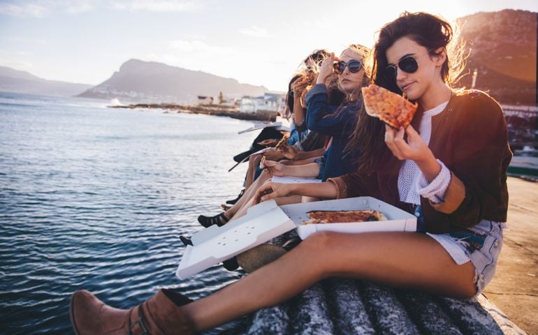 Group of hipster friends eating pizza sitting on the dock at the harbor during a summertime road trip