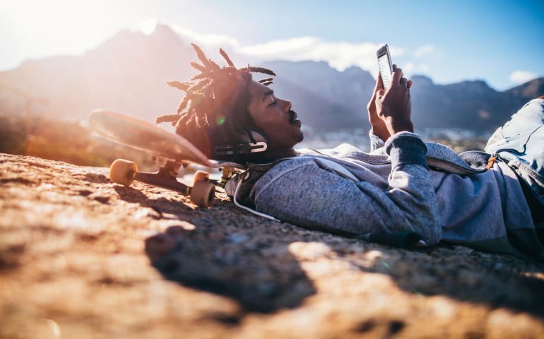 African American skater lying down, listening to music from his smartphone with headphones and resting his head on his skateboard on the rocks of a seaside during sunset
