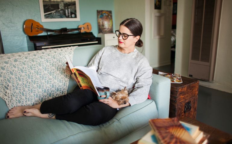 A woman curled up on a blue sofa in her creative home reading a book with her small dog in her lap.