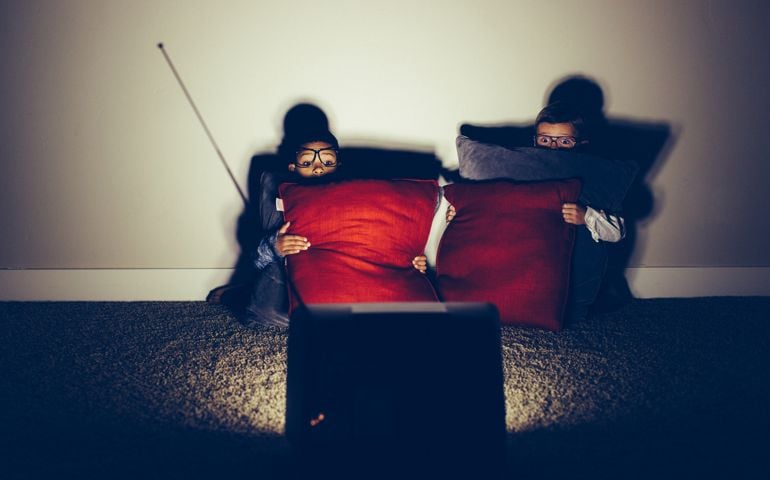 Two young boy nerds sit in a basement late at night on a weekend watching a horror flick. They are both frightened by this scary movie and are hiding behind their pillows while the television glows in the dark.