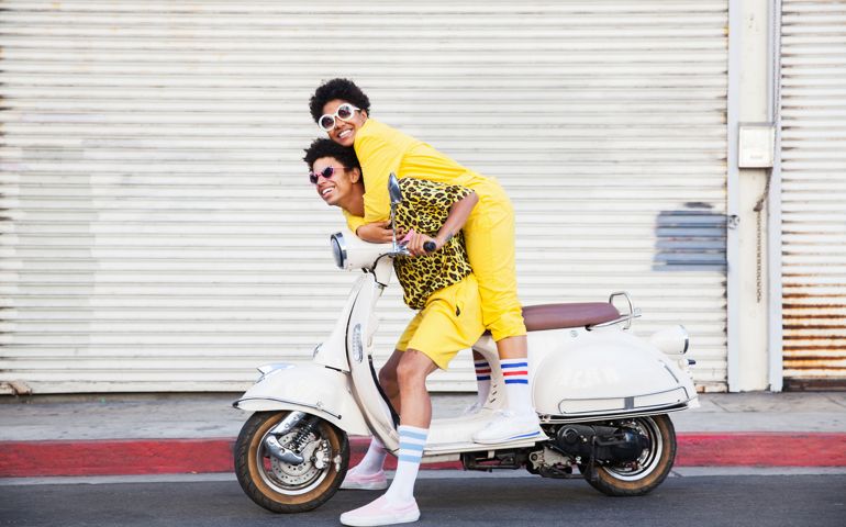 A hip young African American couple wearing bright yellow outfits posing on a scooter on a city street