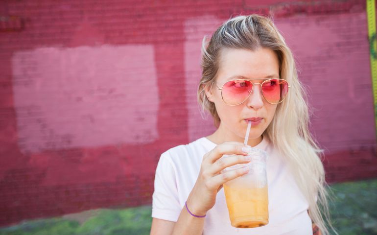 Young blonde woman wearing pink sunglasses drinking juice from a a plastic cup and straw in front of a pink wall