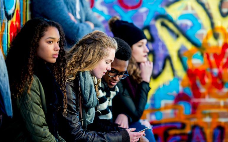 A group of teenagers are sitting in front of a wall covered in graffiti. They are wearing stylish clothes. A boy and girl are looking at a smartphone screen together.