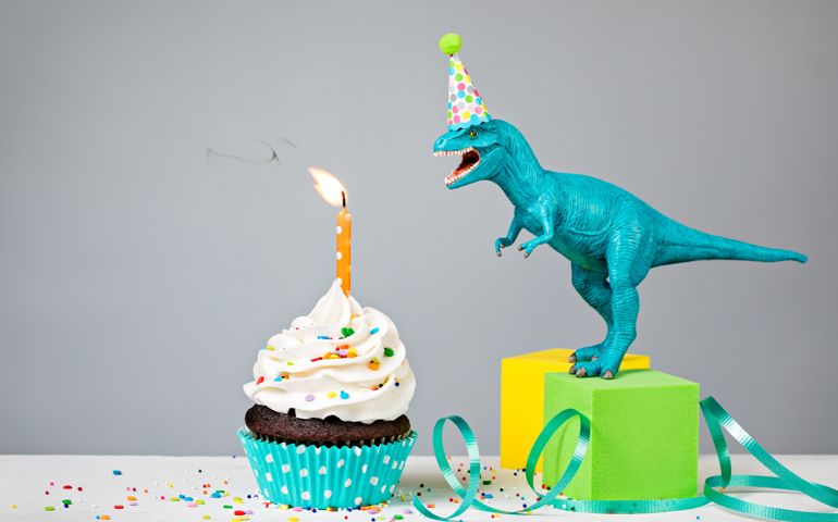 Toy Dinosaur blowing out a Birthday candle with cup cake on a gray background