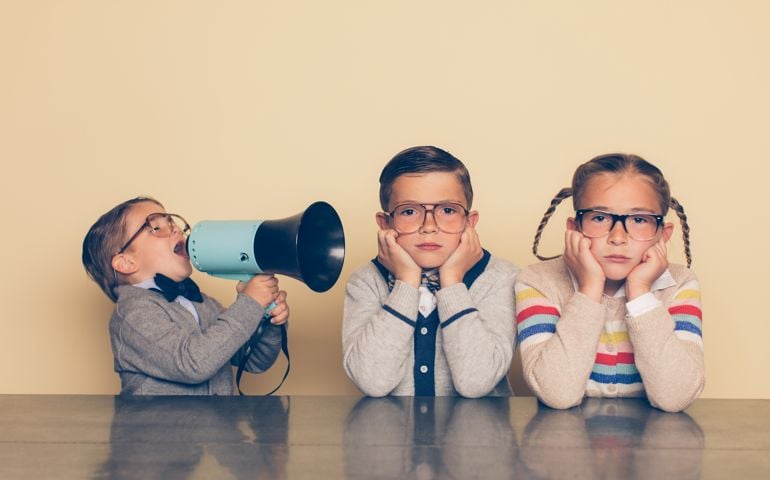 A young boy in glasses and a nerd outfit is shouting at his siblings through a megaphone. The big brother and big sister are looking at the camera with an annoyed look on their faces while plugging their ears. The little brother is bossing his older siblings around.