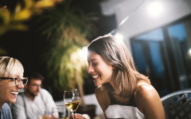 Family sitting and drinking white wine on the terrace at summer night. Mother and daughter laughing so hard. Mom remembered something from the past.