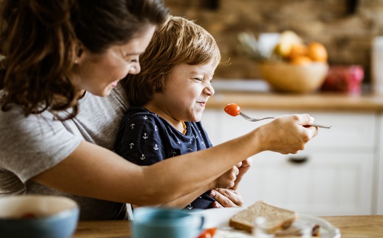 Displeased boy refusing to eat cherry tomato while mother is feeding him at dining table.