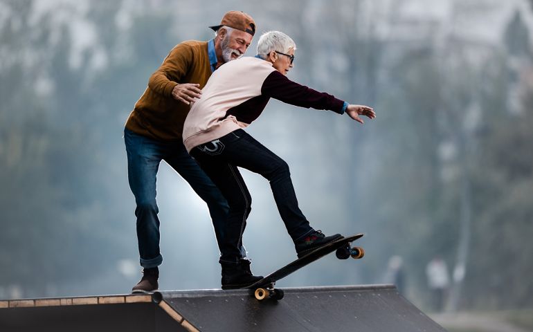 Senior woman riding skateboard on a ramp while her husband is assisting her in a skate park.