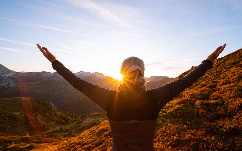 freedom, happiness, closeness to nature, rear view upper body woman with outstretched arms standing high up on alpine mountain pasture enjoying sunset over mountain range in autumn, star shapped sun
