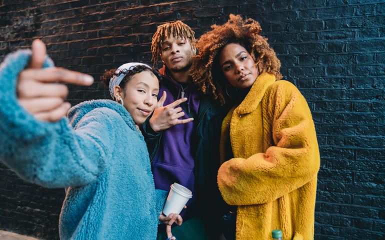 Portrait of three friends against a black brick wall, making gestures in front of the camera. They are wearing colorful and hip clothes.