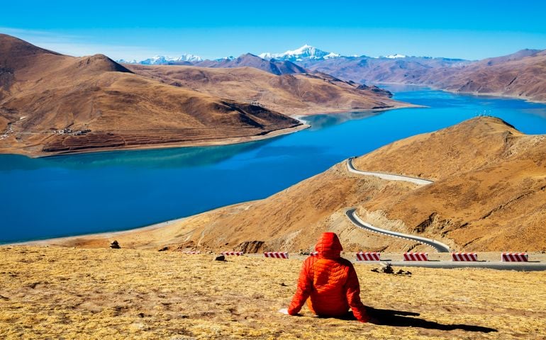 Rear view of female hiker sitting at a height of 4900 m in Tibet. In the background is seen Yamdrok Lake (Yamdrok Yutso in Tibetan dialect) and the Noijinkangsang peak (7206 m) in Himalaya. Yamdrok Lake is situated about 100 km from Lhasa, the capital of Tibet.