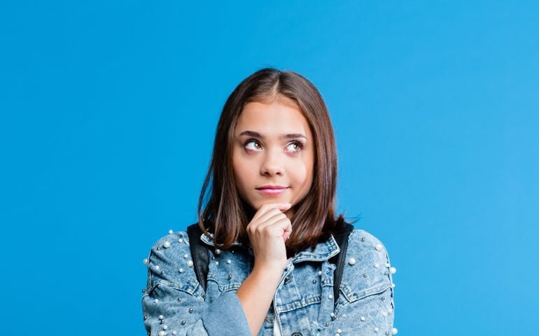 Cute female high school student wearing oversized denim jacket and white t-shirt standing against blue background. Portrait of pensive teenager with hand on chin.