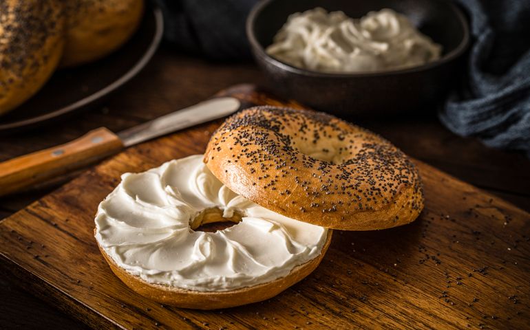 High angle view of an opened poppy seeds bagel spread with cream cheese. The bagel is on a rustic wooden table and it's surrounded by a black plate with two bagels and a bowl with cream cheese. Low key DSLR photo taken with Canon EOS 6D Mark II and Canon EF 100 mm f/ 2.8