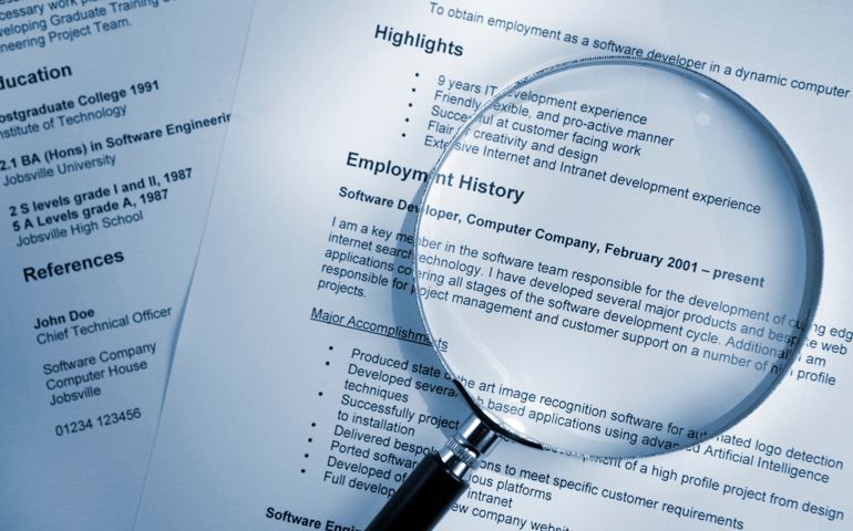 A blue tinted image of a magnifying glass lying on top of several sheets of a printout of resume documents. All information on the resume is fake.