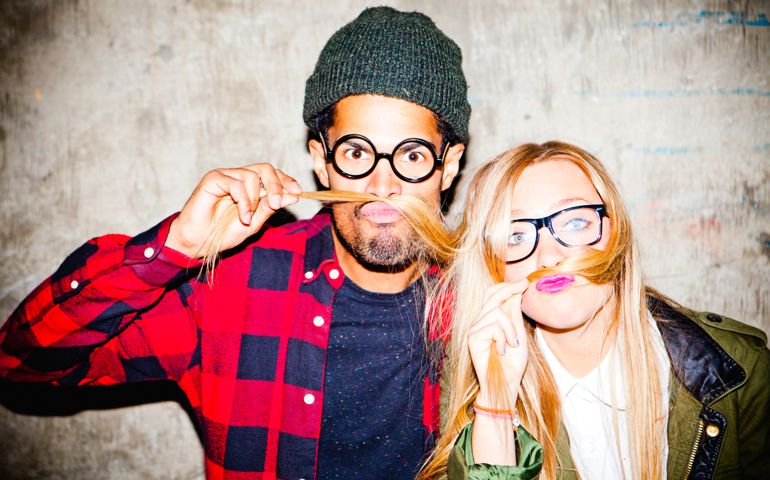 Portrait of a young man and woman pulling funny faces and messing around, standing in front of a rustic background.