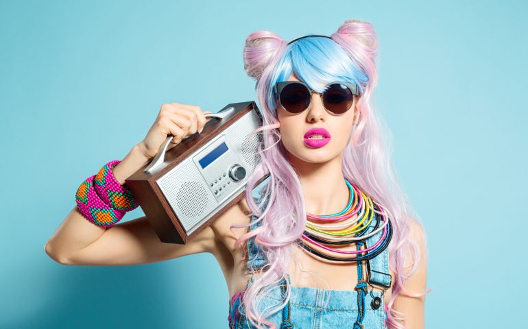 Portrait of sensual manga style blue-pink hair young woman wearing denim coveralls and sunglasses. Standing against blue background, holding radio on her shoulder. Studio shot, one person.