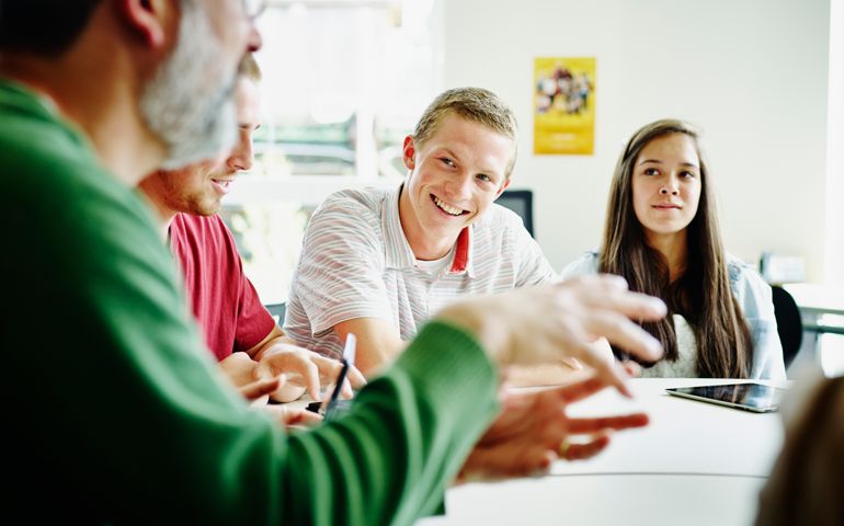 Smiling students sitting at table in high school classroom listening to teacher lecture on project