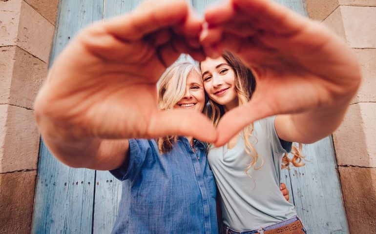 Cheerful grandmother and teenage granddaughter forming a heart shape with their hands in old town