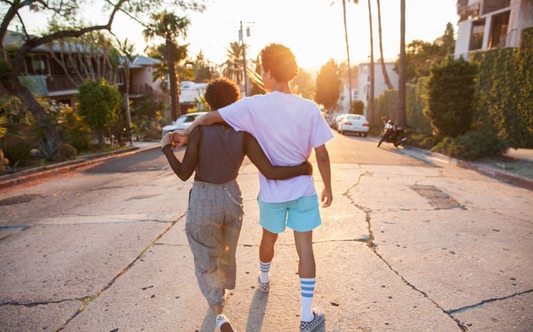 A hip young African American couple with their arms around each other walking down a suburban street at sunset shot from behind