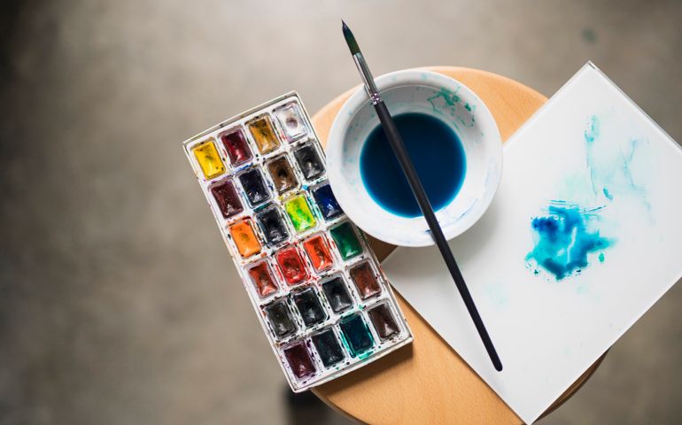 Artist's palette with painting colors, bowl with diluted blue paint and a paintbrush on the chair.