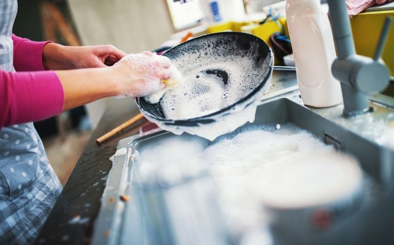 Closeup of unrecognizable woman doing dishes with lots of dirty plates and pots waiting aside. She's cleaning a black non stick cooking pan.