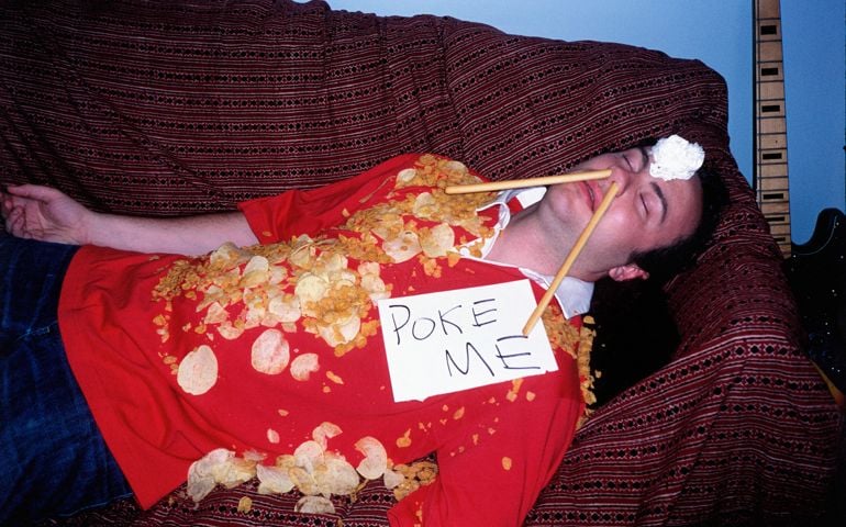 Young man asleep on sofa covered in crisps wearing sign
