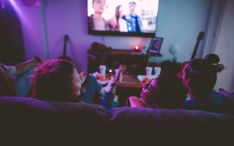 Young multi-ethnic hipster friends relaxing on living room sofa and watching film on TV