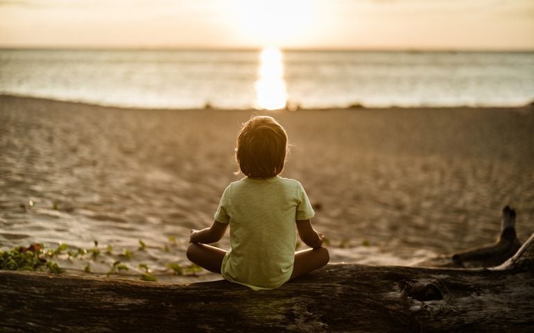 Back view of Zen-like kid practicing Yoga at the beach at sunset.