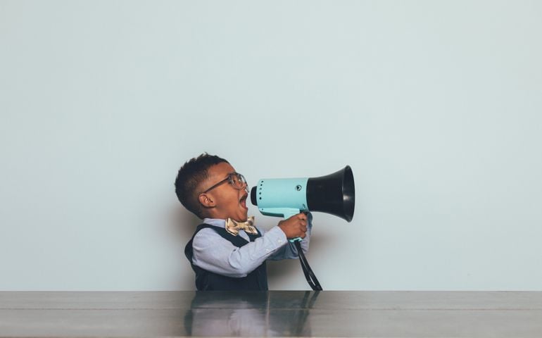 A young nerd boy dressed in bow tie, vest and wearing eyeglasses, holds and yells through a megaphone trying to get someone to listen to him. He sits at a table trying to get his message across to others.