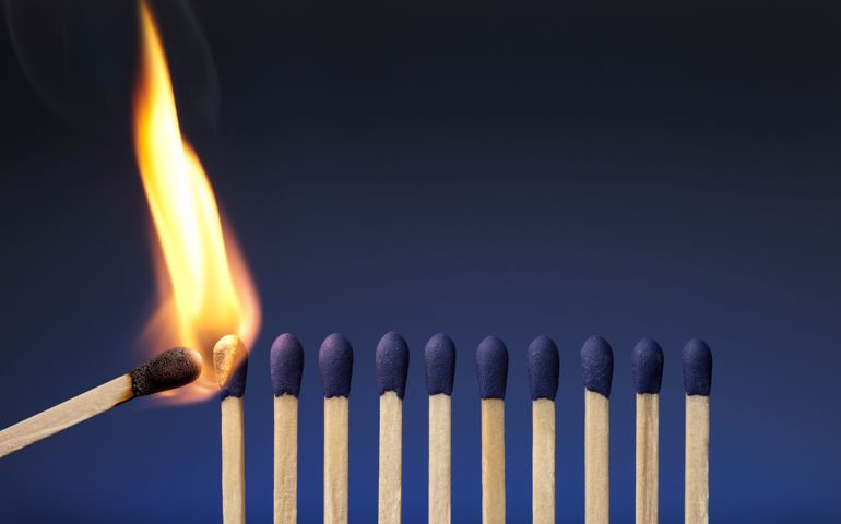 A photograph of blue-tipped matches on a blue background. A single lit match is coming in from the left side of the image and beginning to ignite the leftmost match. The tip of the lit match is black, and the flame is orange. The background of the picture is deep blue at the top and gradually lightens to a brighter blue as it reaches the bottom of the image.