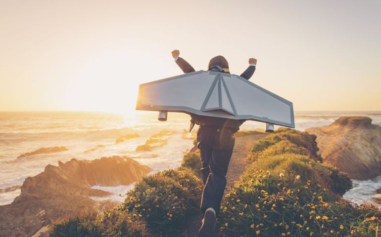 A young boy dressed in business suit and tie wears a homemade jetpack and flying goggles raises his arms in the afternoon sun while running to take off into the air on an outcropping above the surf in Montana de Oro State Park, California. This young entrepreneur is ready to take his new business to new heights.