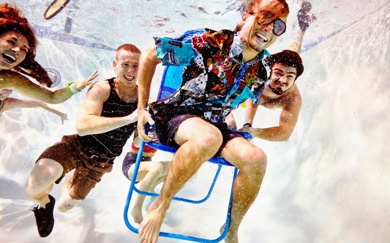 Group of smiling young friends underwater in pool lifting friend in chair
