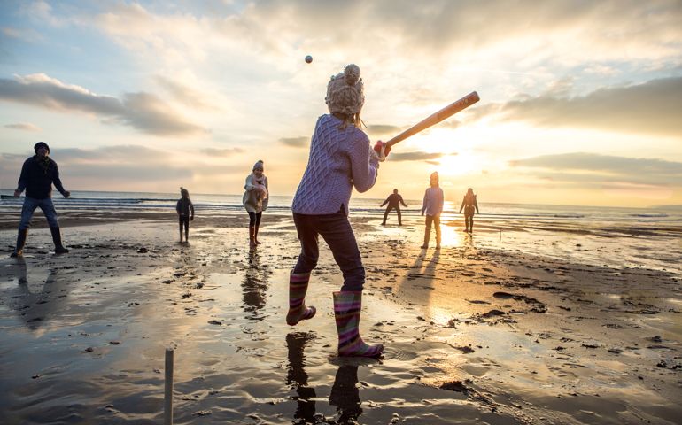 family playing baseball on beach at sunset.