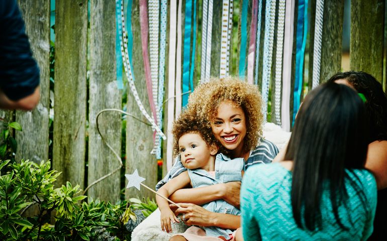 Smiling mother embracing toddler daughter during backyard birthday party with family and friends