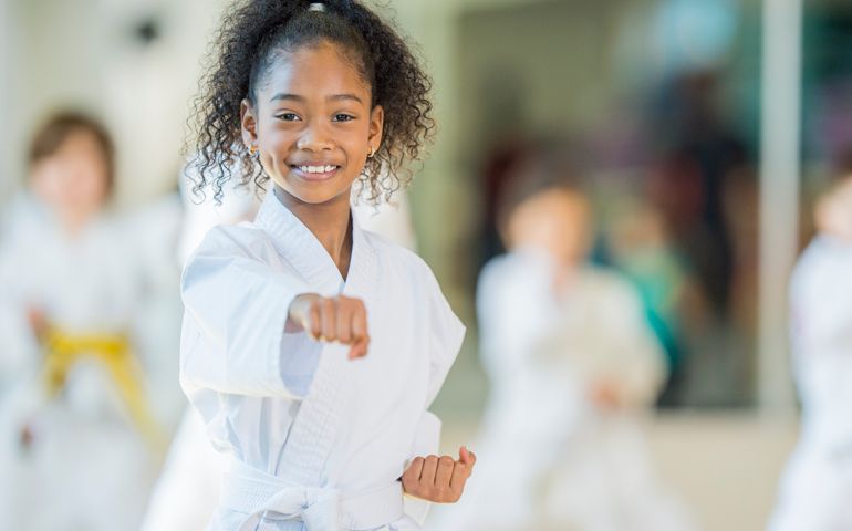 A multi-ethnic group of children are indoors at a Taekwondo academy. They are wearing martial arts clothing. A girl of African descent is punching while smiling at the camera.