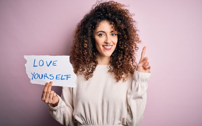 Young beautiful woman with curly hair and piercing holding paper with love yourself message surprised with an idea or question pointing finger with happy face, number one