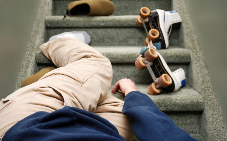 A man trips over roller skates on a staircase in a home. He is not recognizable and is laying head down. Stairs have a muted green colour. Roller skates are old style and white in colour.