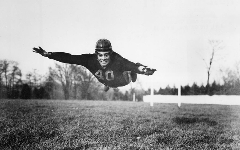 circa 1935: American football player Vince Lombardi (1913 - 1970), wearing a Fordham University uniform and a helmet, diving into the air outdoors in a field.