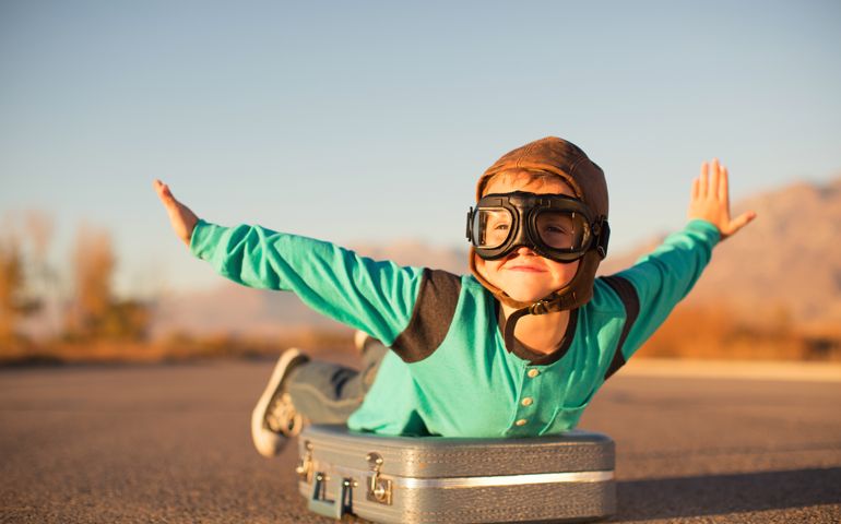 A young boy with outstretched arms lies on top of a suitcase imagining he is flying on an airplane away and traveling to exotic locations. He is wearing a flight cap and goggles and has a large smile on his face. He loves to travel.