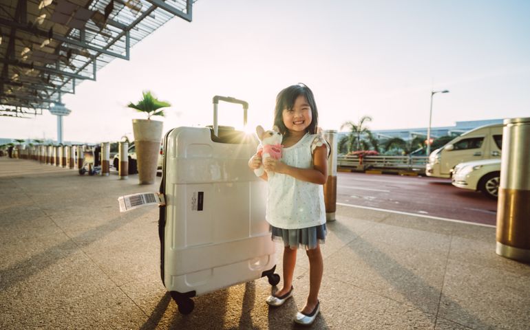 Lovely little girl with huge luggage next to her smiling joyfully while standing in front of hotel with morning sun light behind her.
