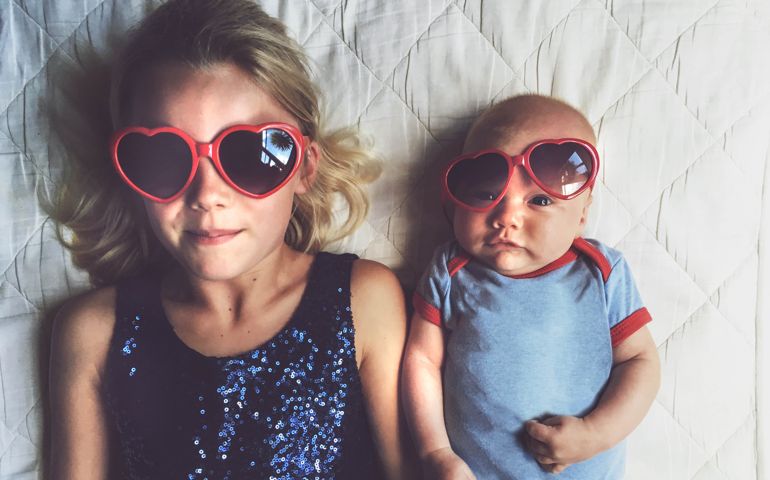 Young girl laying beside baby brother both wearing red love heart glasses