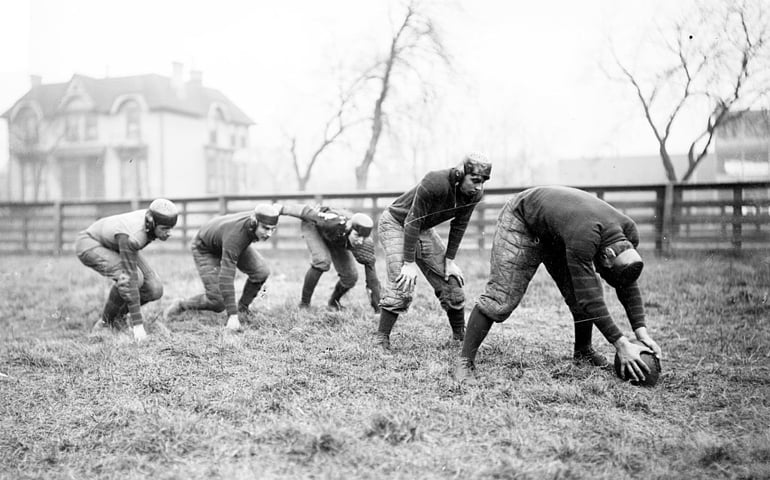 UNITED STATES - CIRCA 1902: Image of three Waller High School football running backs crouching in position behind the center and quarterback on a field in Chicago, Illinois, 1902. A large house is visible in the background. Waller High School (formerly North Division High School) was located at West Armitage Avenue and North Orchard Street in the Lincoln Park community area of Chicago, Illinois. (
