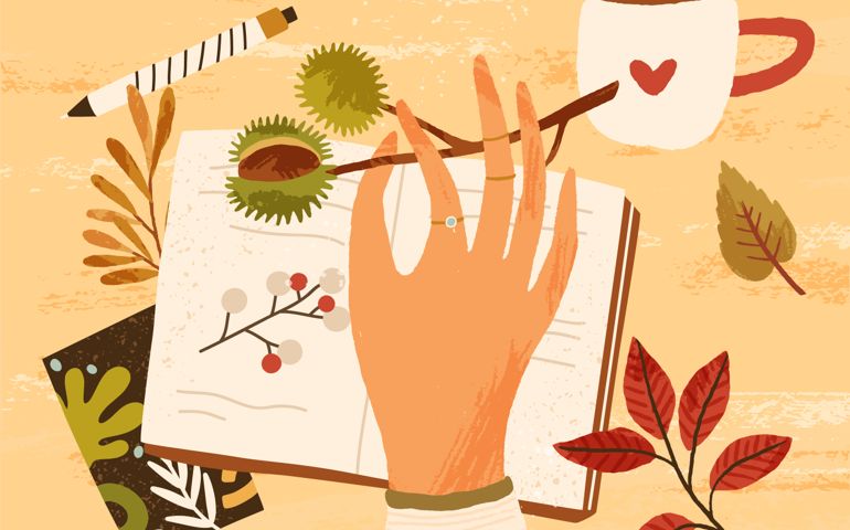 Cozy autumn composition with hand holding branch with chestnut, fall leaves, berries, notebook, pen and a cup of hot beverage