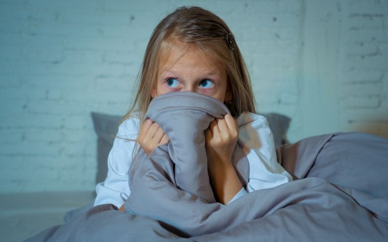 Scared little girl staying sleepless hiding behind the duvet looking horrified in the dark having childhood nightmares in child imagination Sleeping disorders Stress Depression and Insomnia concept.