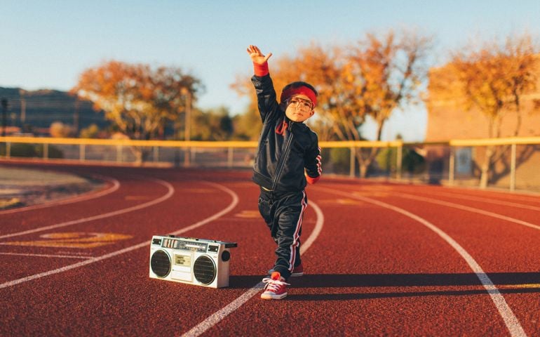 A young nerd boy dressed in a retro track suit and headband stands on the running track with his ghetto blaster ready to live a healthy lifestyle.