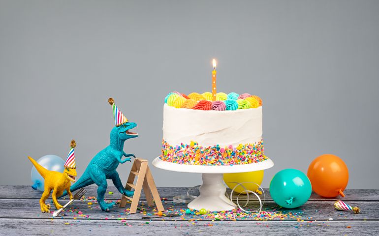 Hungry toy dinosaurs wearing hats and holding forks next to a birthday Cake on a gray background