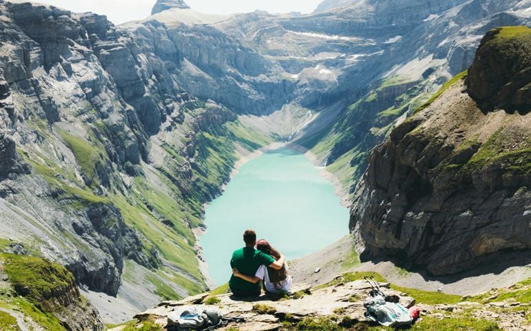 Young woman and man backpackers getting up to the top of the mountains with scenic view of turquoise colored lake in Swiss Alps, embracing loving each other and looking at the beautiful panoramic view in Switzerland
