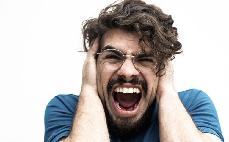 Nervous desperate guy covering ears with hands and shouting loud. Handsome bearded young man in blue casual t-shirt posing isolated over white background. Stress concept